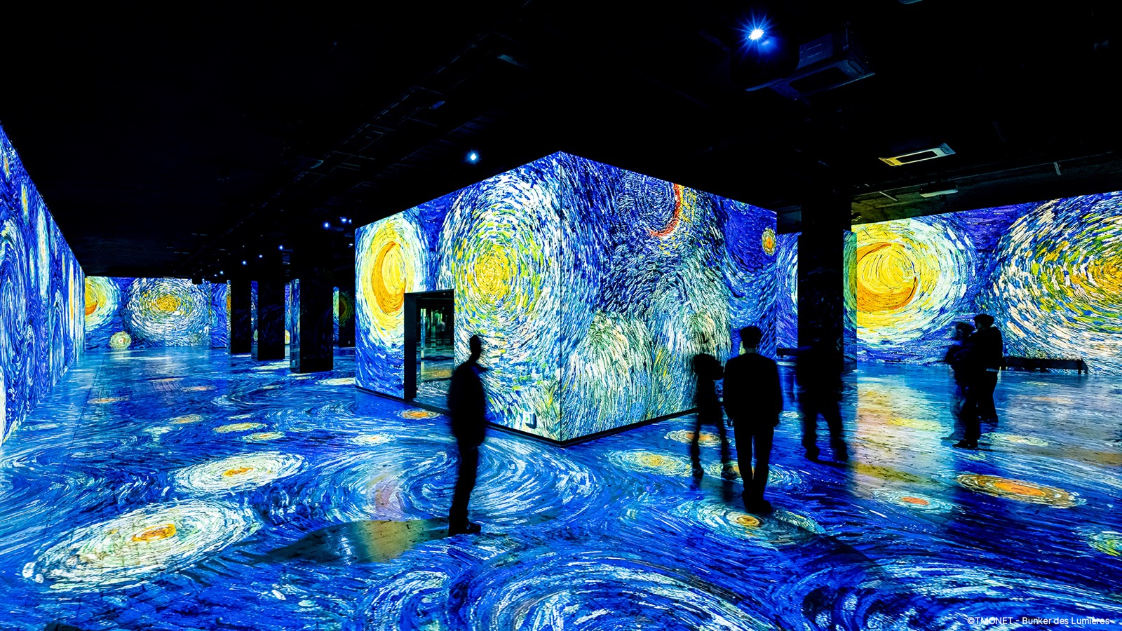 ゴッホの「星月夜」が壁と床の全面に投影された暗い展示空間を、来場者が歩いている没入型メディアアートの場面です。